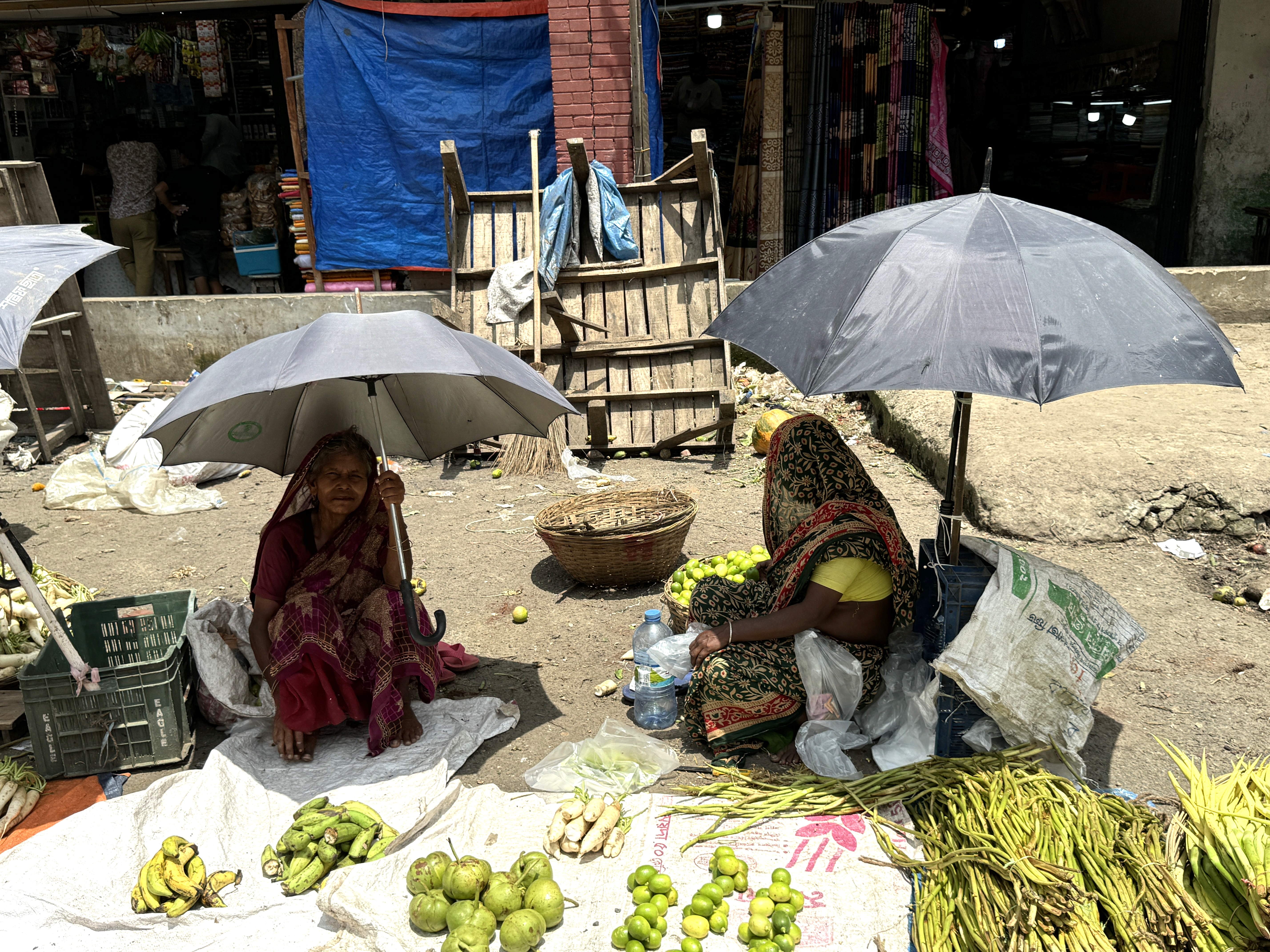 Two migrant women selling vegetables on a hot day in Dhaka’s Karwan Bazar use umbrellas to try to cool themselves in August 2024. Credit: Mosabber Hossain